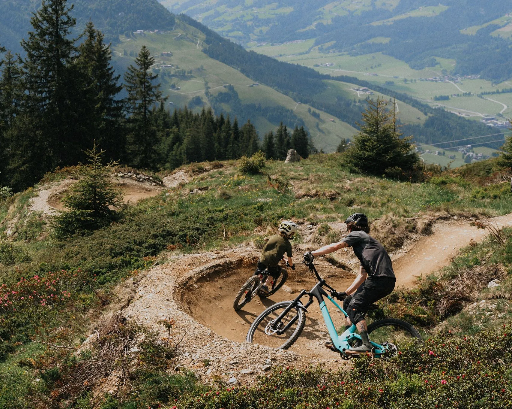 Zwei Mountainbiker fahren eine Kurve auf einem Flowtrail mit Blick auf die umliegenden Berge.